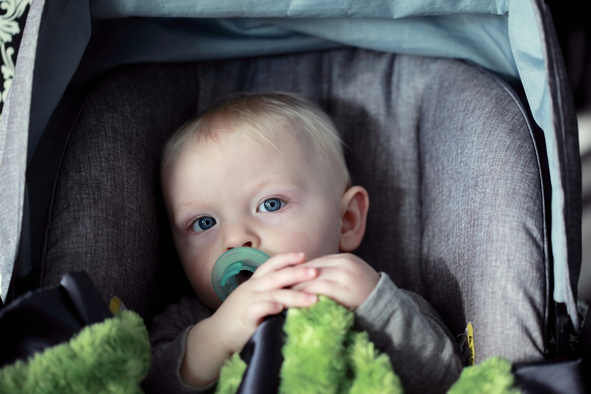 A baby with light hair and blue eyes sits in a gray fabric car seat or stroller, wearing a gray long-sleeved garment and sucking on a green pacifier. A fuzzy green blanket is visible in the foreground, partially obscuring the lower portion of the image.