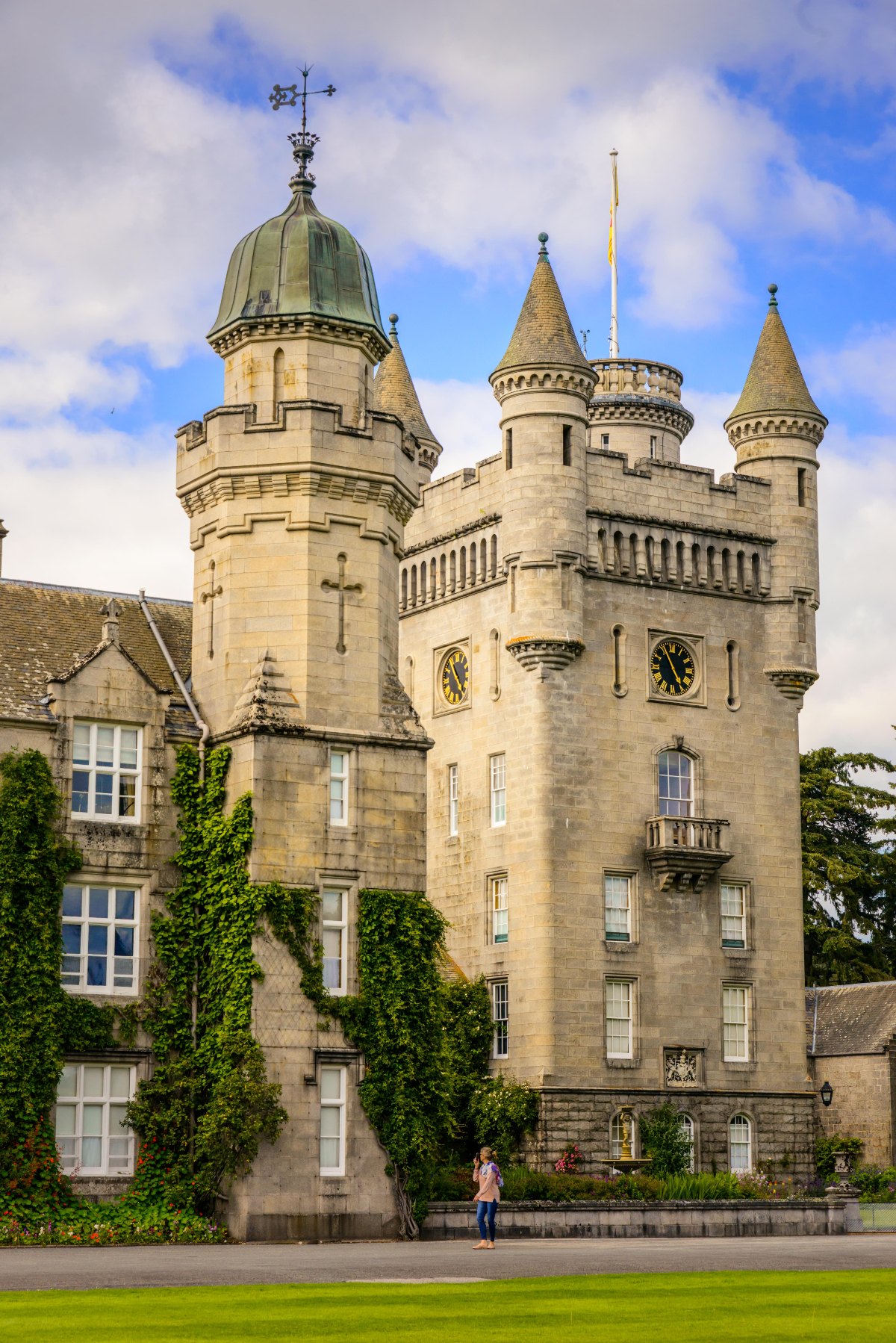 The pale granite towers of Balmoral Castle rise against a partly cloudy sky, featuring multiple conical turrets, a green copper-domed clock tower on the left, and a second clock face on the central tower, with ivy climbing the lower walls of the adjoining wing. A single visitor stands on the path below, providing scale against the castle's imposing facade, with a manicured lawn in the foreground.