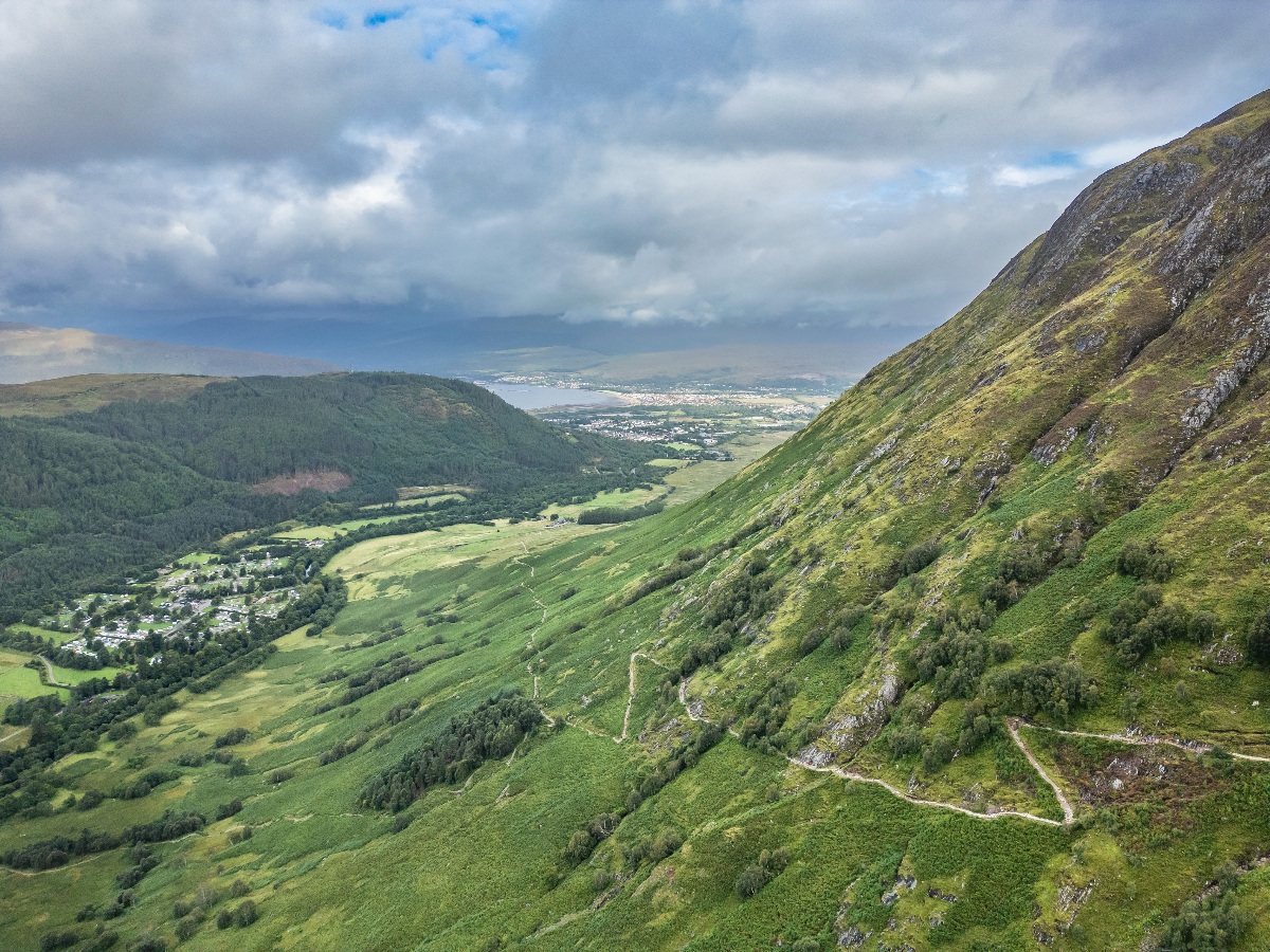 A steep, rocky mountainside dominates the right of the frame, with a switchback hiking trail zigzagging down its green slope toward a broad valley below. A small town and a glimmer of water — likely a loch — are visible in the distance, flanked by forested hills under a heavy, cloud-filled sky.
