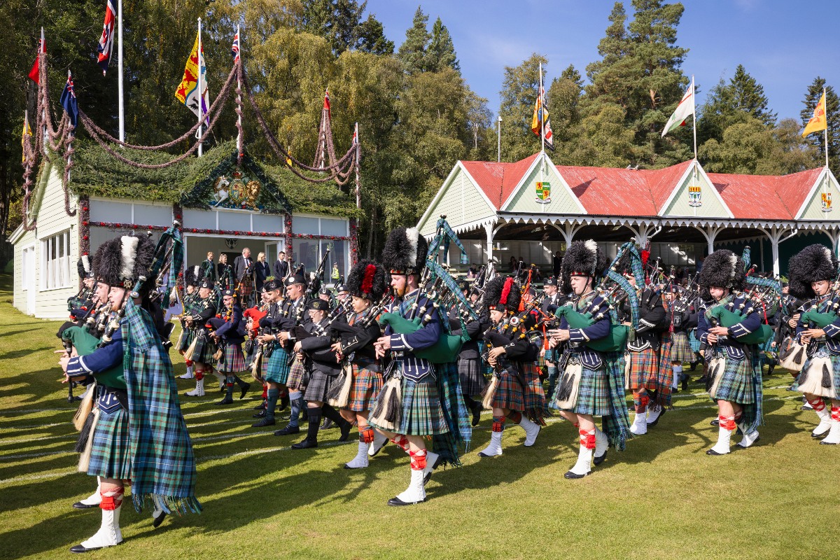 A large massed pipe band marches across a grass field in full Highland dress — kilts in various tartans, green and navy jackets, and tall black bearskin hats — each player carrying bagpipes. Behind them, white pavilion buildings decorated with garlands, crests, and multiple national flags line the grounds, with tall pine trees visible against a clear blue sky.