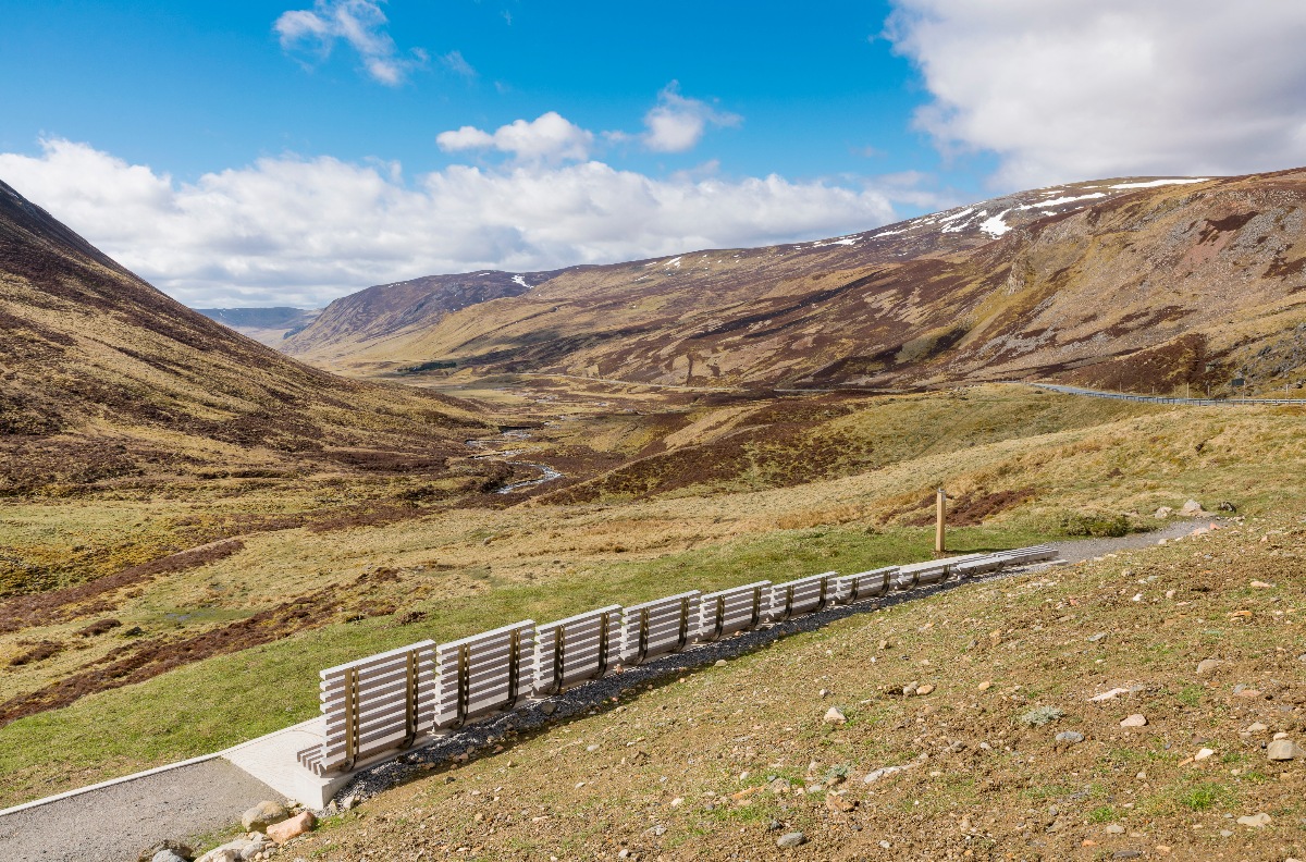 A long, low slatted viewing bench sits on a paved overlook above a wide Highland glen, with a narrow river threading through the valley floor far below and a winding road visible on the right hillside. The surrounding moorland hills are covered in brown and amber heather, with patches of snow remaining on the higher peaks under a bright blue sky with scattered clouds.