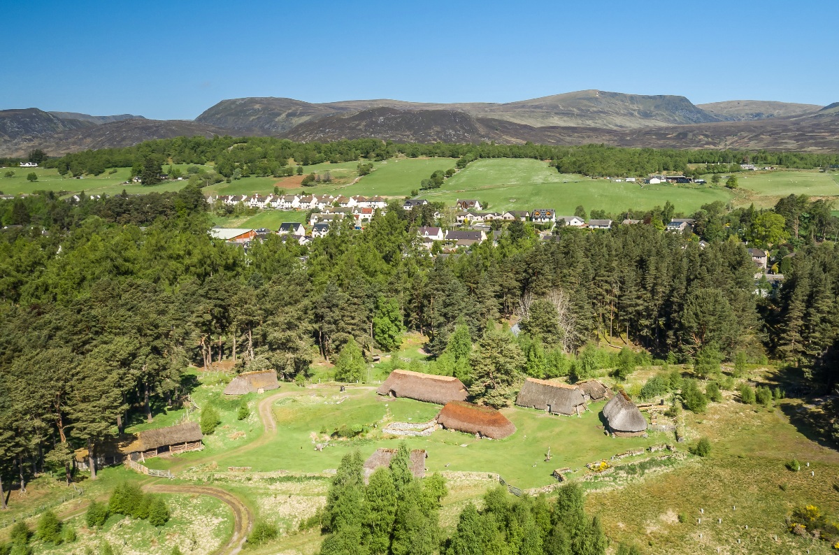 An aerial view shows a cluster of reconstructed thatched-roof Highland buildings — longhouses and roundhouses in various sizes — arranged on a green clearing amid tall pine and mixed woodland. A modern village with white-painted houses is visible through the trees beyond, with open green fields and bare moorland hills stretching to the horizon under a clear blue sky.