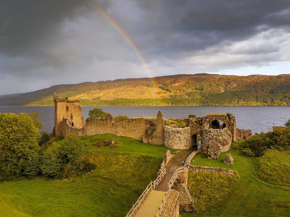The ruins of Urquhart Castle — including a standing tower, crumbling curtain walls, and a round tower gatehouse — sit on a grassy promontory directly on the shore of Loch Ness, with a paved visitor path and white railings leading into the site. A rainbow arcs through a dramatic storm sky above the opposite bank, where autumn-toned hills and forest are briefly lit by a break in the clouds.