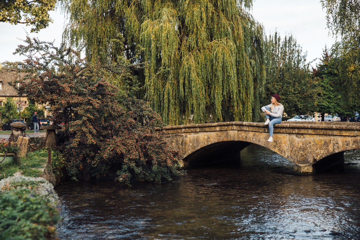 A woman wearing a burgundy hat and jeans sits on the edge of a low, honey-colored stone arch bridge spanning a flowing river, with a large weeping willow and a red-berried shrub rising behind her. Cotswold-stone buildings and additional trees are visible in the background, suggesting a village setting.