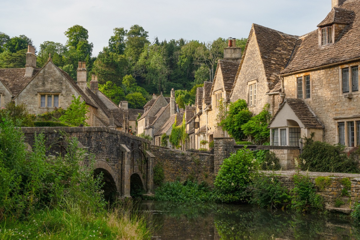 A medieval stone arch bridge spans a calm stream flanked by lush green vegetation, with a row of traditional Cotswold-stone cottages extending along the bank into the background. Wooded hills rise behind the honey-colored village buildings, which feature steeply pitched stone-tile roofs, multiple chimneys, and climbing greenery along their facades.