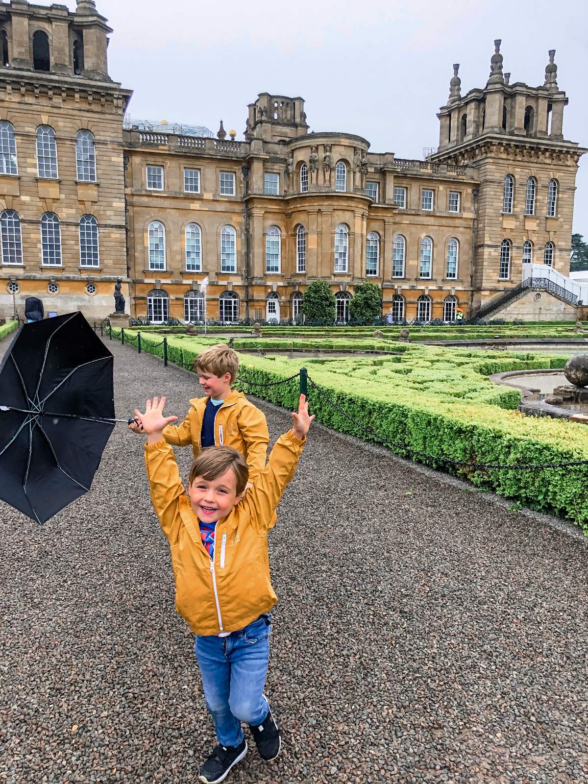 Two children wearing matching yellow rain jackets stand on a gravel path in front of a large baroque palace, with one child raising both arms and smiling at the camera while a black umbrella rests on the ground nearby. Formal parterre gardens with neatly clipped low hedges extend behind them toward the ornate stone façade of the palace under an overcast sky.