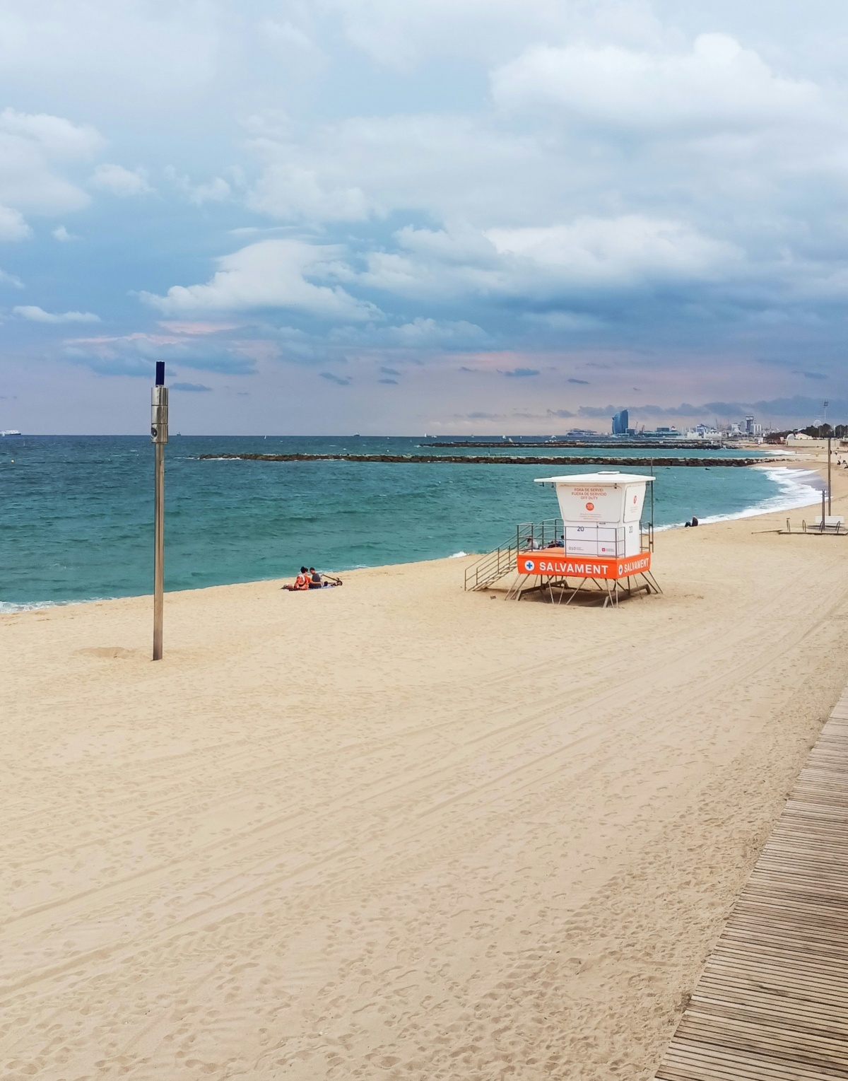 A quiet, mostly empty sandy beach with an off-duty orange and white "Salvament" lifeguard tower, a wooden boardwalk along the right edge, and rock breakwaters extending into the turquoise sea under a cloudy sky. In the distance, a small group sits on the sand near the water's edge, with Barcelona's city skyline faintly visible along the curve of the coast.