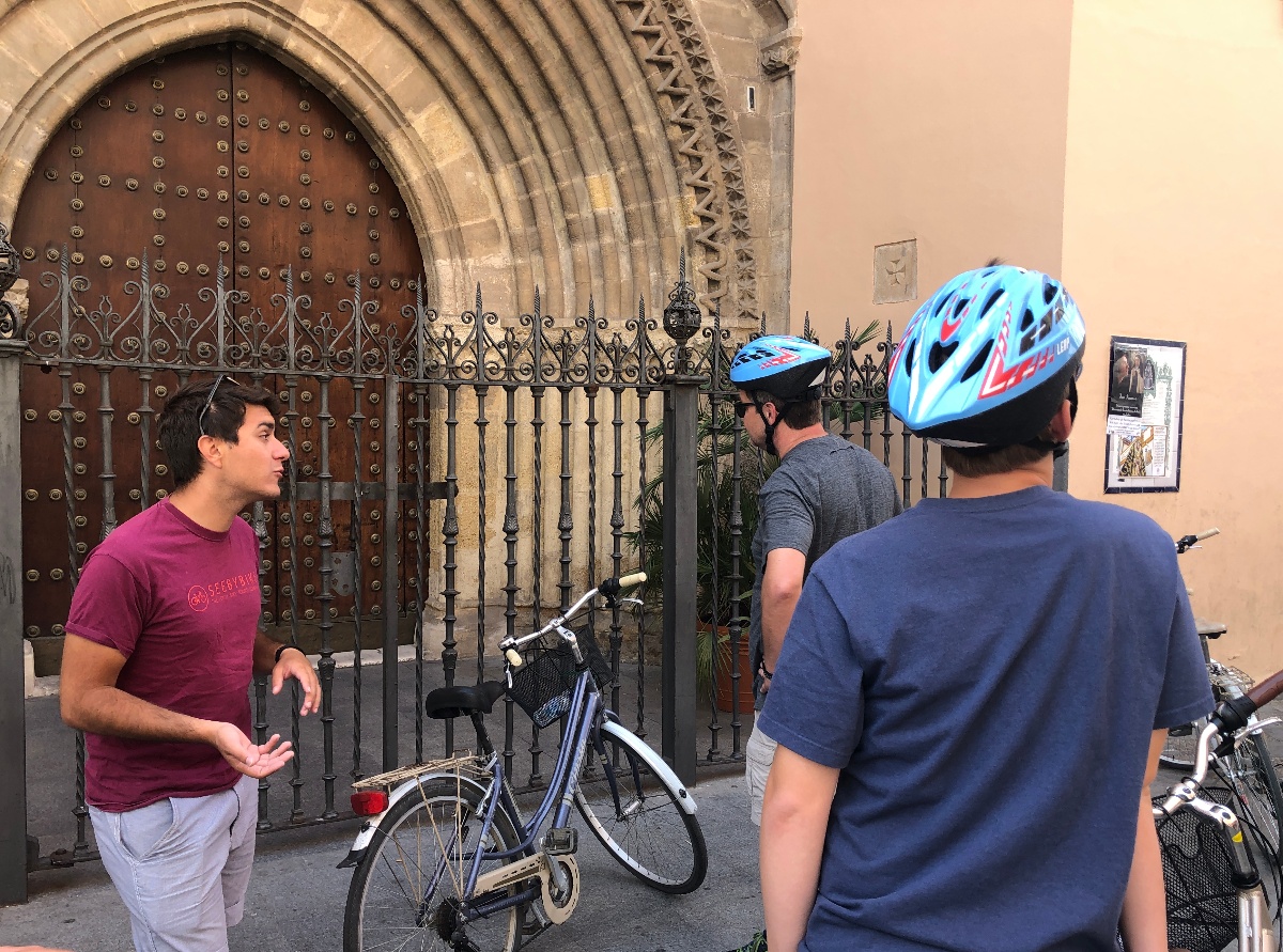 A tour guide in a red t-shirt gestures while speaking to two bicycle tour participants wearing helmets, with bikes parked in front of an ornate iron fence and a large arched wooden door set into a stone Gothic-style church entrance. The group stands on a narrow street beside a warm ochre-colored building, with decorative ironwork and studded wooden doors visible in the background.