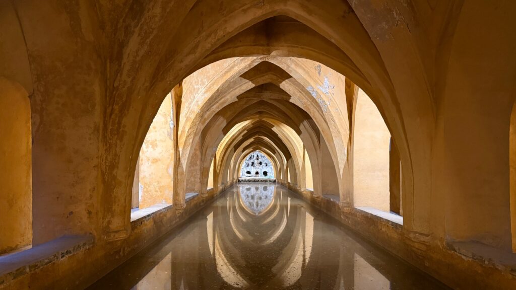 A long underground vaulted passage in Seville's Real Alcázar features a symmetrical series of pointed Gothic arches in warm golden stone, receding toward a decorative stone opening at the far end. A shallow water channel runs the length of the floor, perfectly reflecting the arched ceiling above and creating a mirror image that extends the sense of depth.