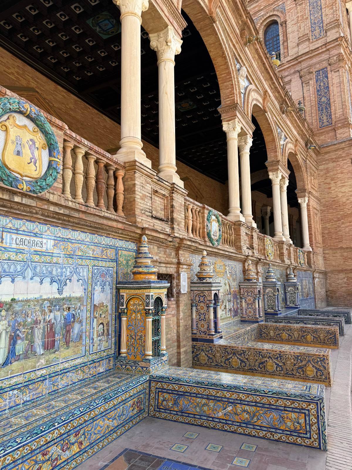 A curved arcade at Seville's Plaza de España features ornate hand-painted azulejo tile benches and panels depicting Spanish provinces, with intricate blue, gold, and green geometric and figurative designs covering every surface. Carved stone columns with a decorated wooden ceiling run along the upper gallery above the tiled alcoves, with the warm terracotta brick facade continuing in the background.