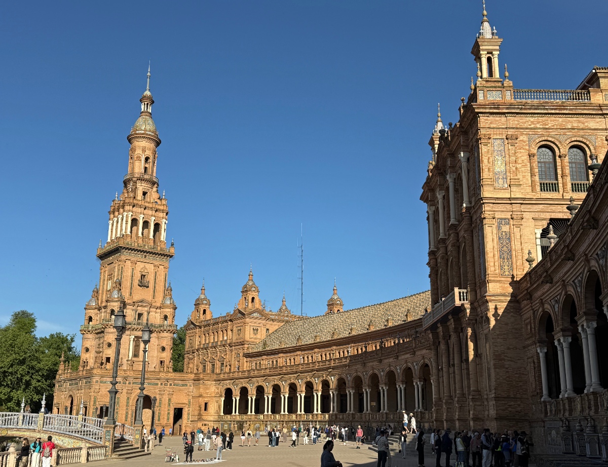 The sweeping terracotta brick facade of Seville's Plaza de España curves across the frame, with a tall ornate tower rising on the left, a colonnaded arcade at ground level, and decorative turrets and tiled rooflines extending across the background under a deep blue sky. Crowds of visitors fill the open plaza in the foreground, with a tile-decorated bridge and ornamental lampposts visible along the left edge.