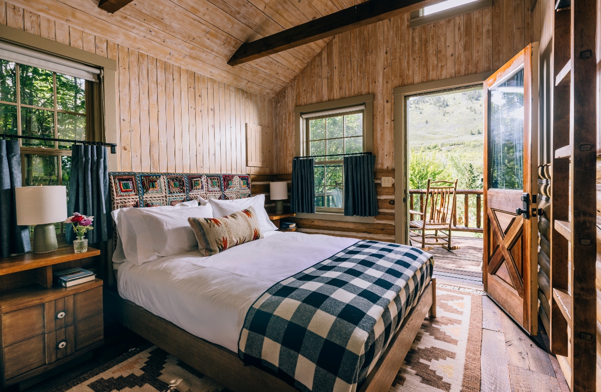 A rustic guest room features light wood paneled walls and vaulted ceiling with exposed dark beams, a bed dressed in white linens with a black and white buffalo check throw and colorful patterned pillows. The room opens through glass doors to a private deck with mountain views, and includes dark curtained windows, simple wood furnishings, and a rocking chair visible on the outdoor space.