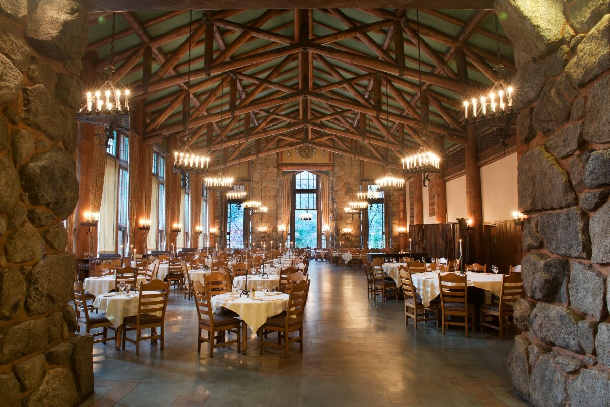 The dining room features massive granite stone pillars framing the space, exposed timber trusses forming an arched ceiling, and multiple wrought iron chandeliers with candle-style lights illuminating the hall. Round dining tables with white tablecloths and wooden chairs are arranged throughout the room, while tall windows and wall sconces provide additional lighting against the stone and wood architecture.