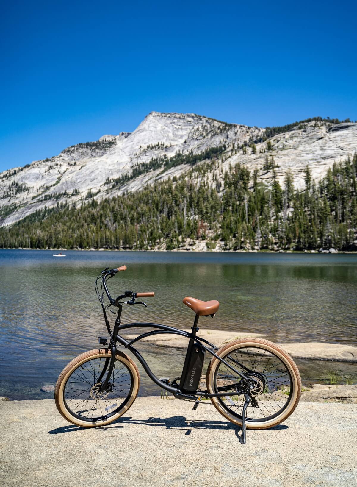 A black electric bicycle with tan leather seat, handlebar grips, and tan-walled tires stands on granite pavement beside a calm alpine lake reflecting the surrounding landscape. Behind the lake, dense evergreen forest covers the lower slopes of a large granite dome formation rising beneath a clear blue sky.