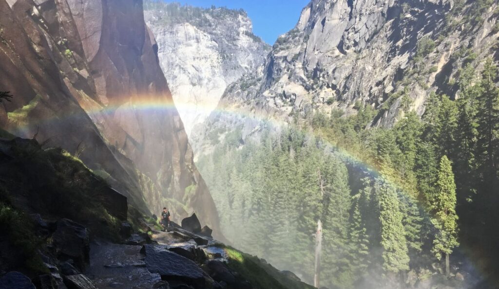 A hiker ascends wet stone steps carved into granite along a narrow canyon, with a powerful waterfall creating dense mist and multiple rainbows visible in the spray. Steep granite cliff walls rise on both sides, covered with evergreen conifers, while sunlight illuminates the waterfall mist against a clear blue sky.
