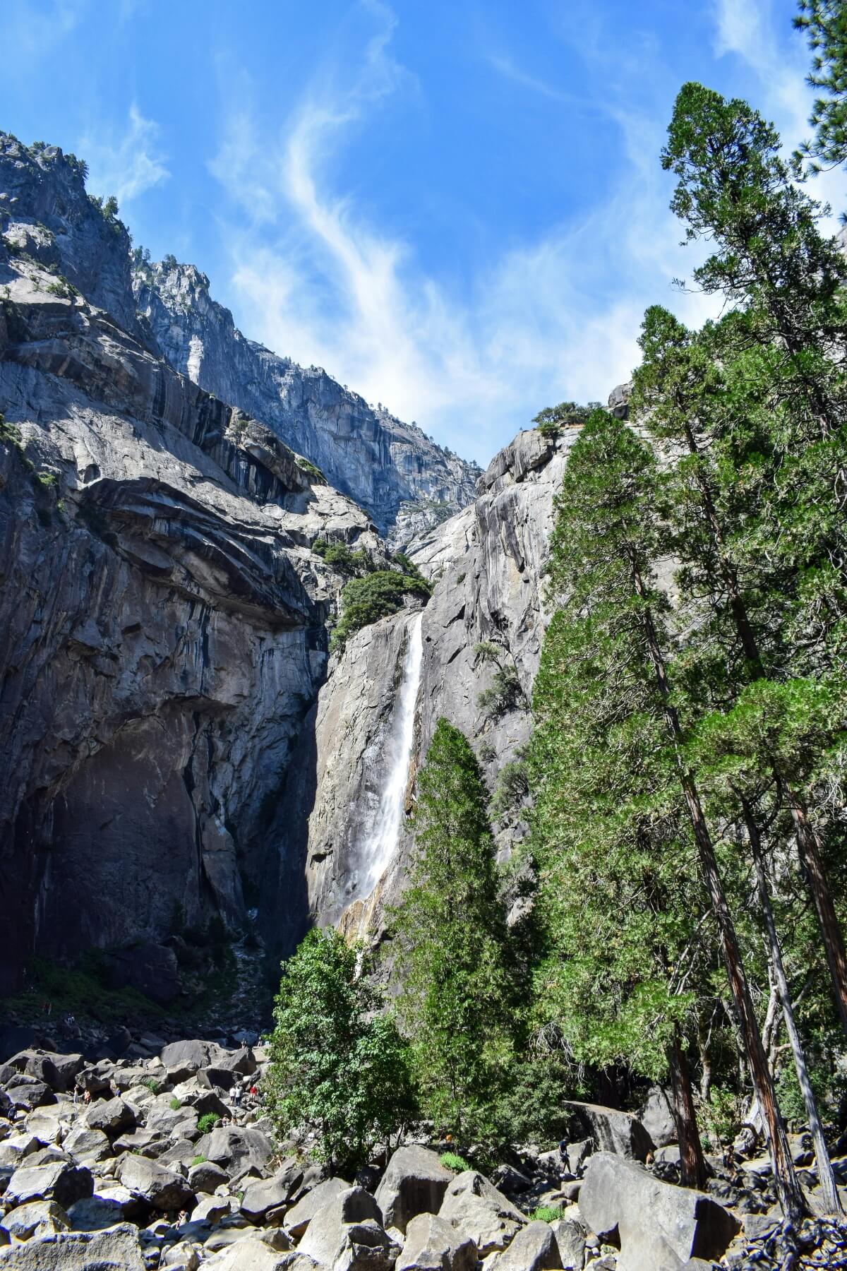 Lower Yosemite Falls drops down a narrow granite cliff face between steep canyon walls, with mist visible at the base where the water meets a boulder field. Visitors gather among large granite rocks at the waterfall's base, surrounded by deciduous trees and evergreen conifers growing along the canyon floor and slopes beneath a blue sky with wispy clouds.