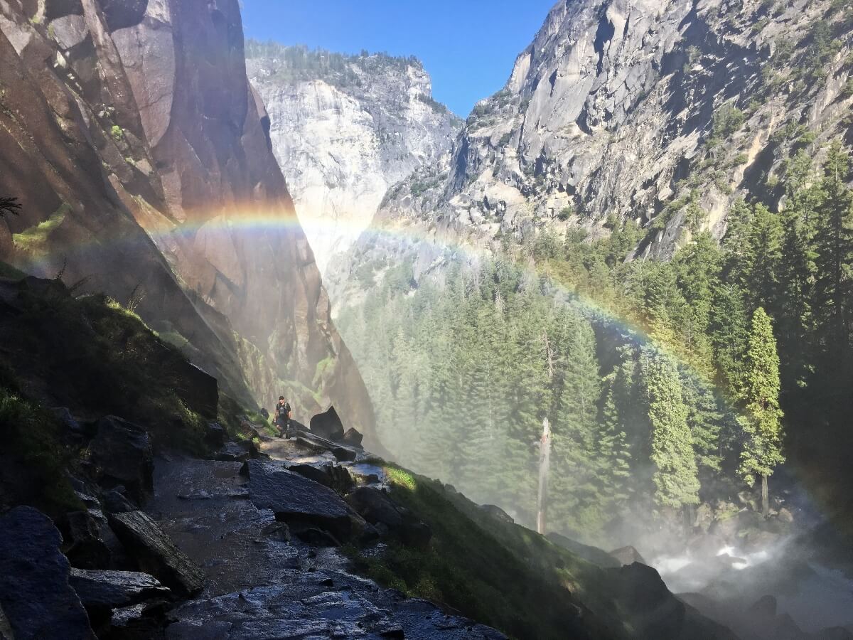 A hiker ascends wet stone steps carved into granite along a narrow canyon, with a powerful waterfall creating dense mist and multiple rainbows visible in the spray. Steep granite cliff walls rise on both sides, covered with evergreen conifers, while sunlight illuminates the waterfall mist against a clear blue sky.