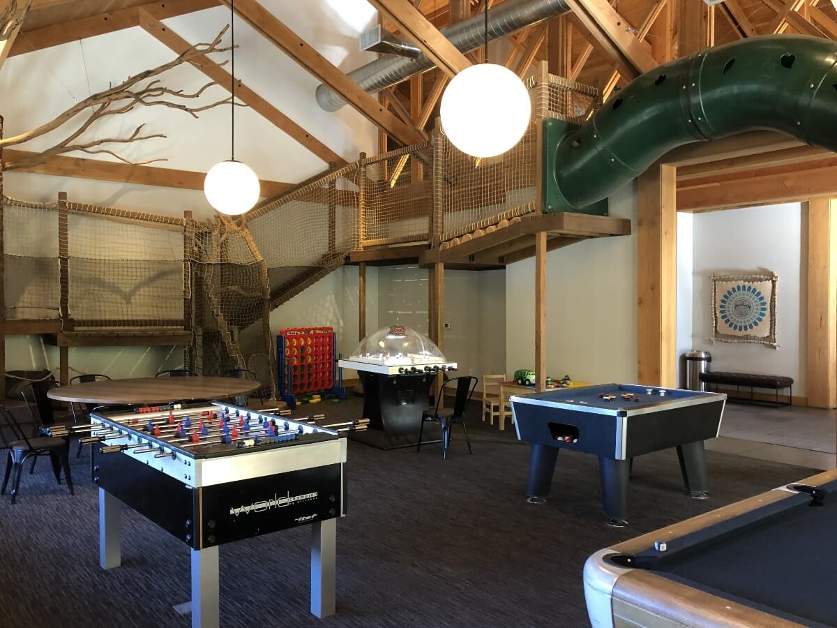 A recreation room features high vaulted ceilings with exposed timber beams, large spherical pendant lights, and a multi-level wooden climbing structure with rope netting and a green tube slide emerging from the upper level. The space includes game tables such as foosball, air hockey, and pool on dark flooring, with natural branch installations and wood-paneled walls throughout the room.