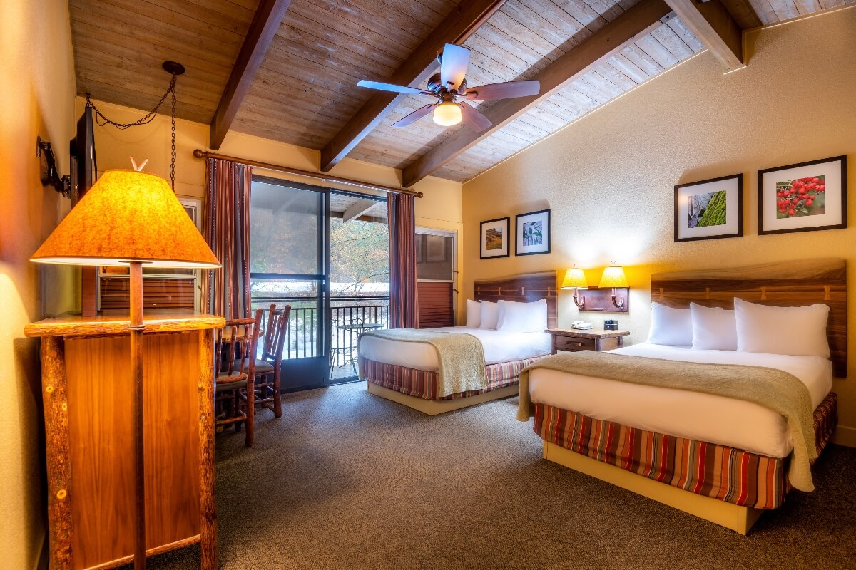 A hotel room features two beds with white linens and striped platform bases in earth tones, positioned beneath vaulted wood-planked ceilings with exposed beams and a ceiling fan. The room includes a rustic wooden table with a large amber lamp, framed nature photography on the walls, and a sliding glass door opening to a balcony with views of trees and natural surroundings.