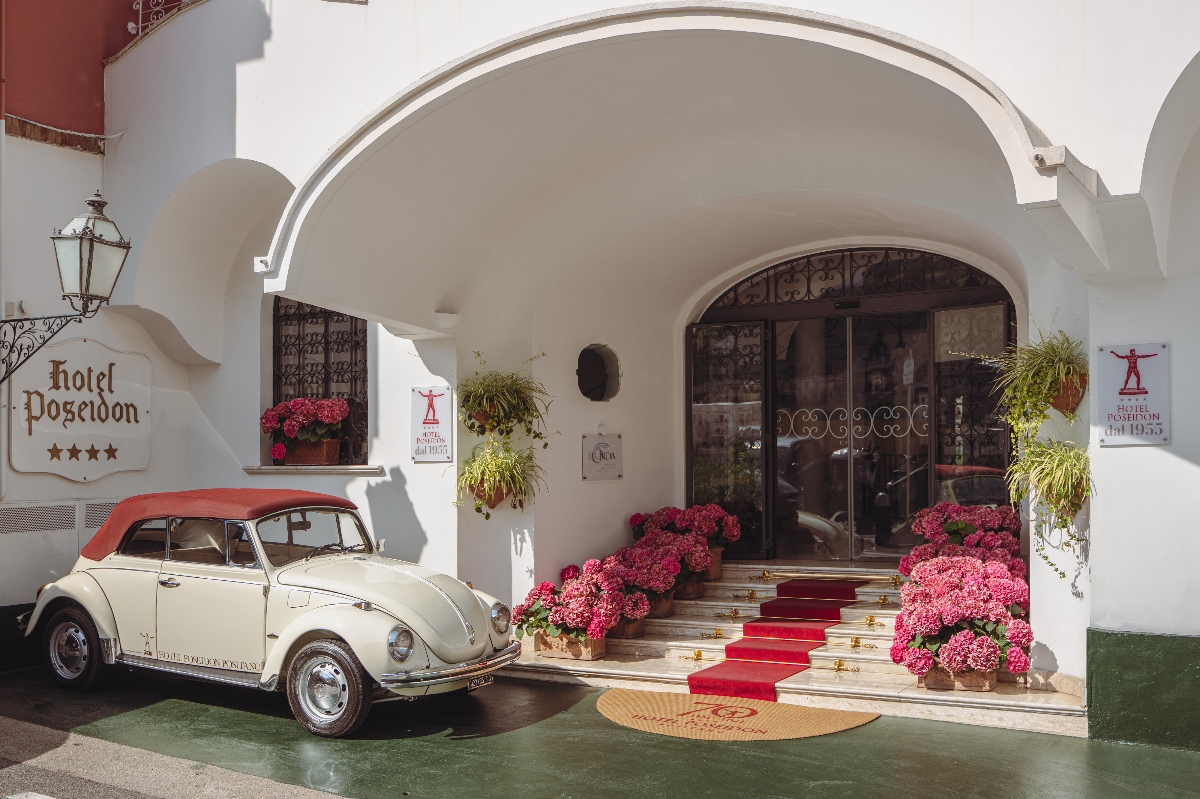 A vintage cream-colored Volkswagen Beetle convertible with a red top is parked in front of a white stucco building with arched architecture and a "hotel Poseidon" sign indicating four stars. Stone steps with a red carpet runner lead to glass entrance doors flanked by large pink hydrangeas in planters, with hanging plants suspended from the arched portico above.