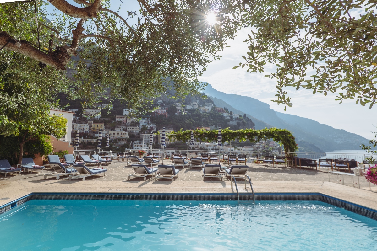 A swimming pool with blue water sits on a terrace with lounge chairs arranged on the deck, framed by olive tree branches in the foreground. Beyond the pool, white buildings of a coastal town rise up a mountainside, with mountains visible in the distance under a bright sky.