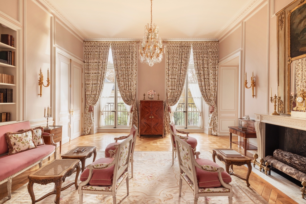 Salon-style living room of the Suite Necker at Airelles Château de Versailles, featuring Louis XVI–style carved armchairs with rose silk upholstery, matching floral toile draperies, herringbone parquet floors, and a crystal chandelier. Floor-to-ceiling paneled walls in blush pink frame tall French doors overlooking garden greenery, with gilt sconces, a marble fireplace, and period antiques completing the 18th-century interior.