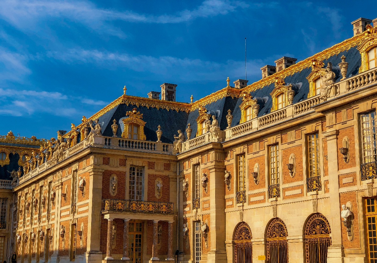 The ornate facade of the Palace of Versailles is bathed in warm golden light, its red brick and cream stone exterior detailed with white marble busts, wrought iron balconies, and elaborate sculptural reliefs. The steeply pitched slate mansard roofline is trimmed with gilded ornamentation and punctuated by decorative dormer windows, stone urns, and classical statuary set against a vivid blue sky. The close-up angle emphasizes the layered depth of the architecture, from the arched ground-floor doorways to the gilded cornices crowning the roofline above.
