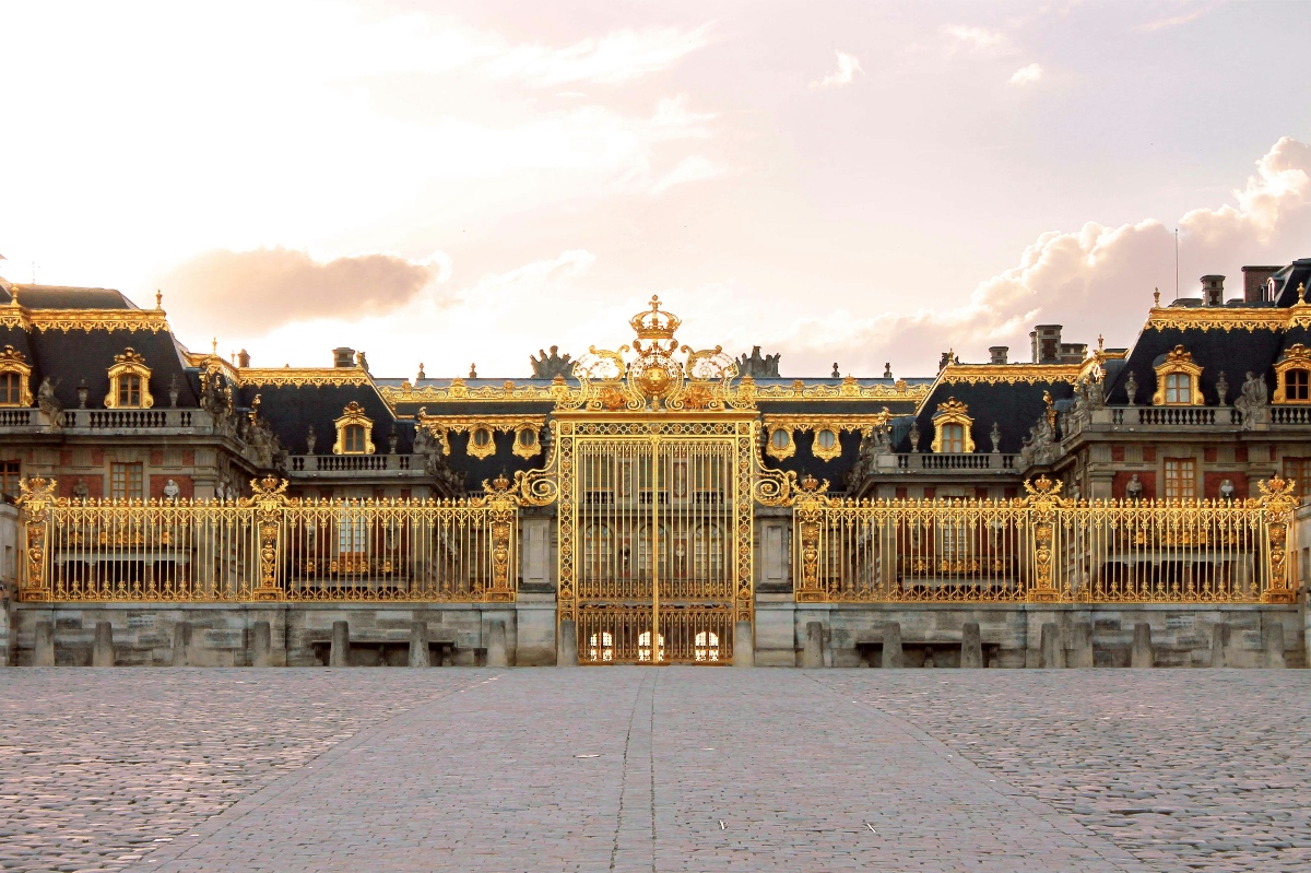 The gilded Royal Gates of the Palace of Versailles stretch across the Cour Royale, their ornate wrought iron panels and elaborate gold scrollwork gleaming against a soft, cloud-streaked sky. A towering central gate is crowned with a royal coat of arms and decorative flourishes, flanked symmetrically by lower fence sections and the palace's distinctive dark mansard rooflines trimmed in gold. The cobblestone forecourt stretches toward the camera in the foreground, empty and wide.