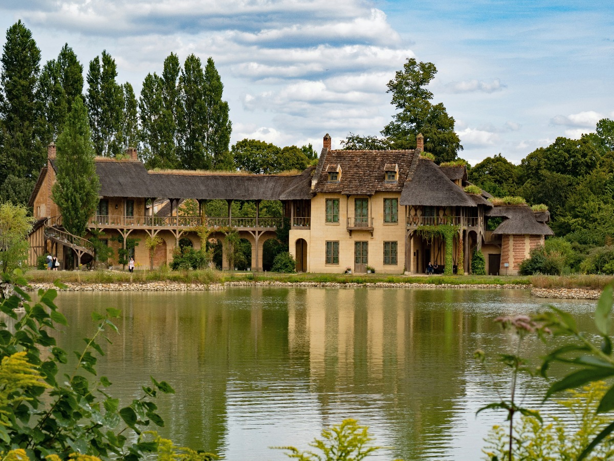 A cluster of rustic Norman-style buildings with thatched and tiled roofs lines the bank of a still pond at the Queen's Hamlet in Versailles, their warm ochre and half-timbered facades reflected in the calm water below. Wooden balconies, climbing vines, and stone archways give the structures a deliberately weathered, pastoral character, set against a backdrop of tall poplars and dense woodland. A handful of visitors are visible along the water's edge on the left, providing a sense of the buildings' scale.