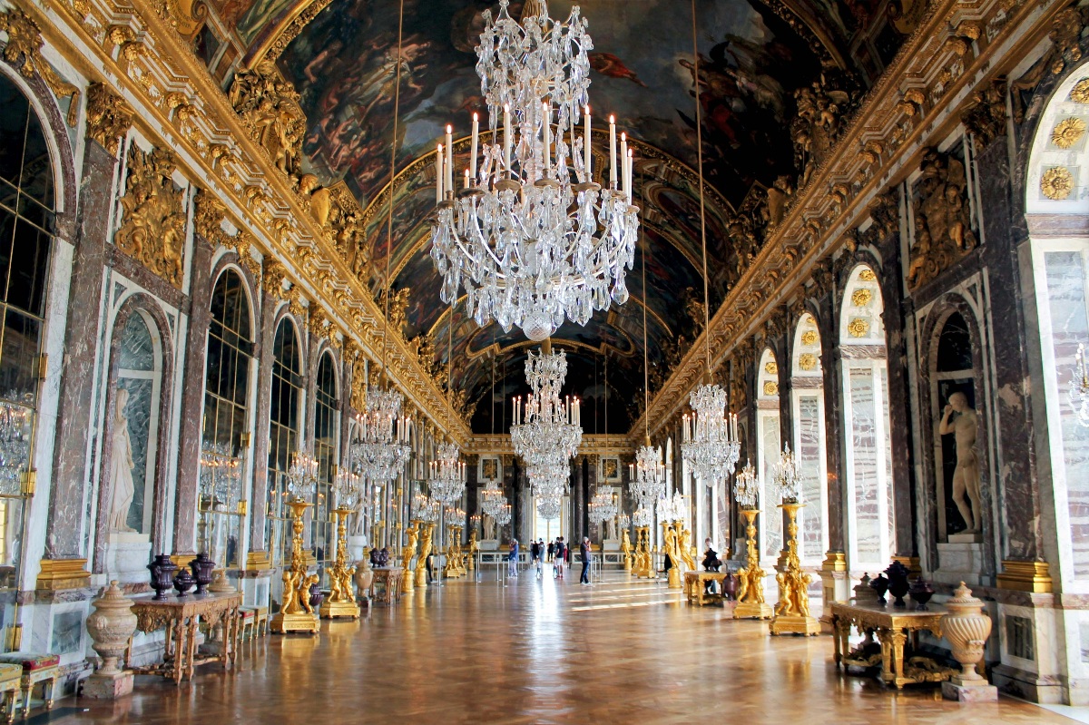 The Hall of Mirrors at the Palace of Versailles stretches into the distance, its vaulted ceiling covered in large-scale painted murals framed by gilded molding and interspersed with ornate medallions. A row of towering crystal chandeliers runs the length of the gallery, flanked on one side by arched windows and on the other by mirrored panels, all set against walls of deep brown marble with gold detailing. The polished parquet floor reflects the light, and a small number of visitors can be seen in the far distance, giving a sense of the hall's vast scale.