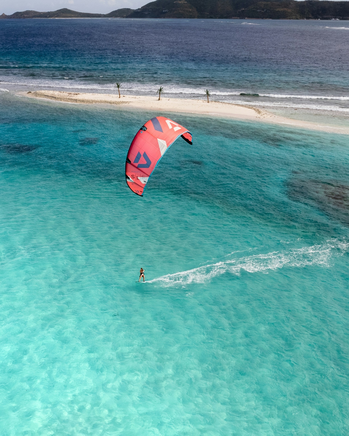 A kitesurfer with a red and white geometric-patterned kite glides across shallow turquoise water, leaving a wake trail behind. In the background, a narrow white sandbar with three small palm trees extends into the water, with dark mountainous islands visible on the horizon.