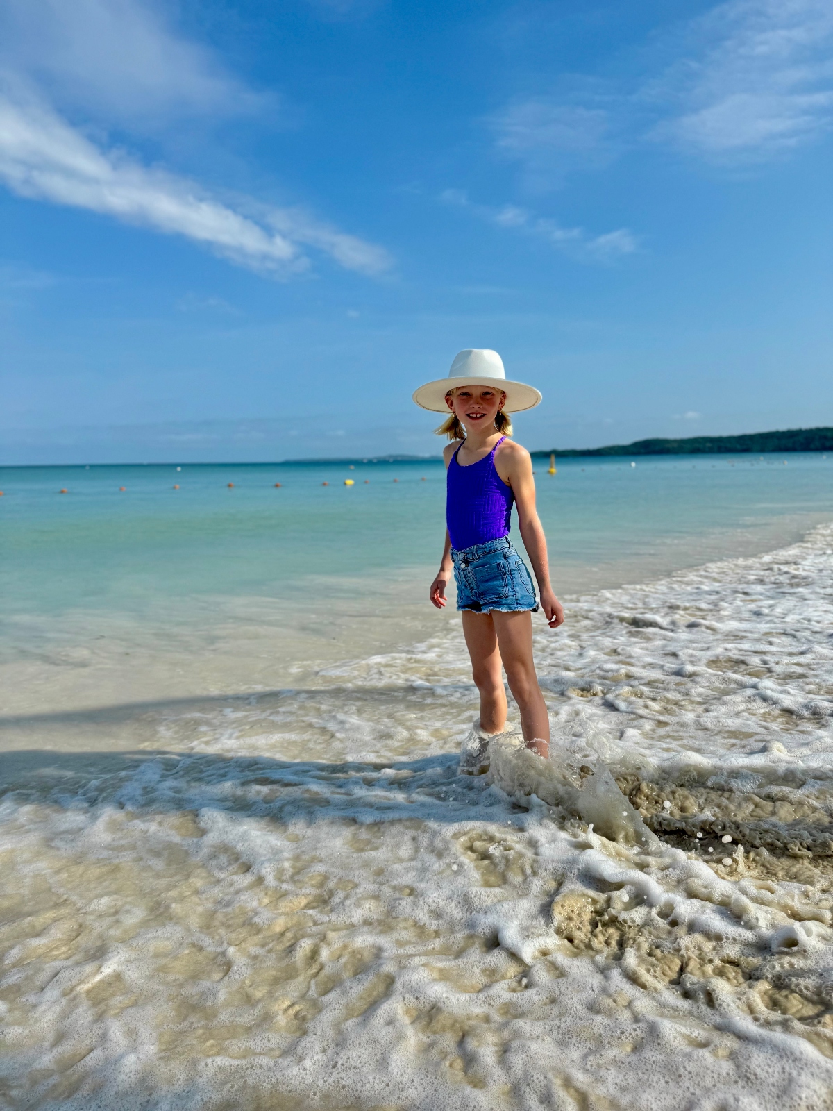 A young girl wearing a white wide-brimmed hat, purple swimsuit, and denim shorts stands in shallow turquoise water as gentle waves wash around her feet. The calm Caribbean sea stretches to the horizon under a blue sky with wispy clouds, with a distant coastline visible in the background.