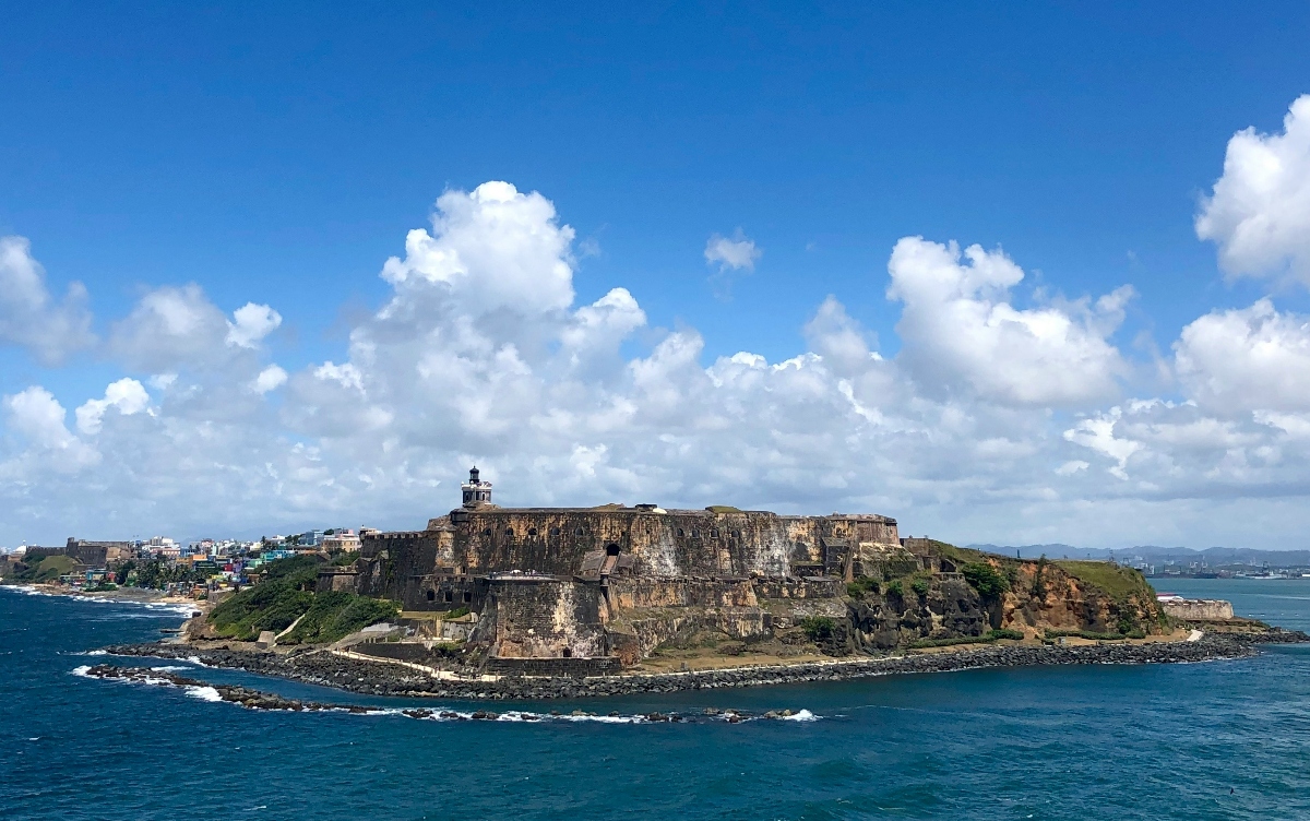 A historic stone fortress with weathered walls sits on a rocky peninsula jutting into turquoise ocean water, with a lighthouse structure atop its ramparts. Colorful buildings are visible on the coastline behind the fort, and white clouds dot the blue sky above the coastal landscape.