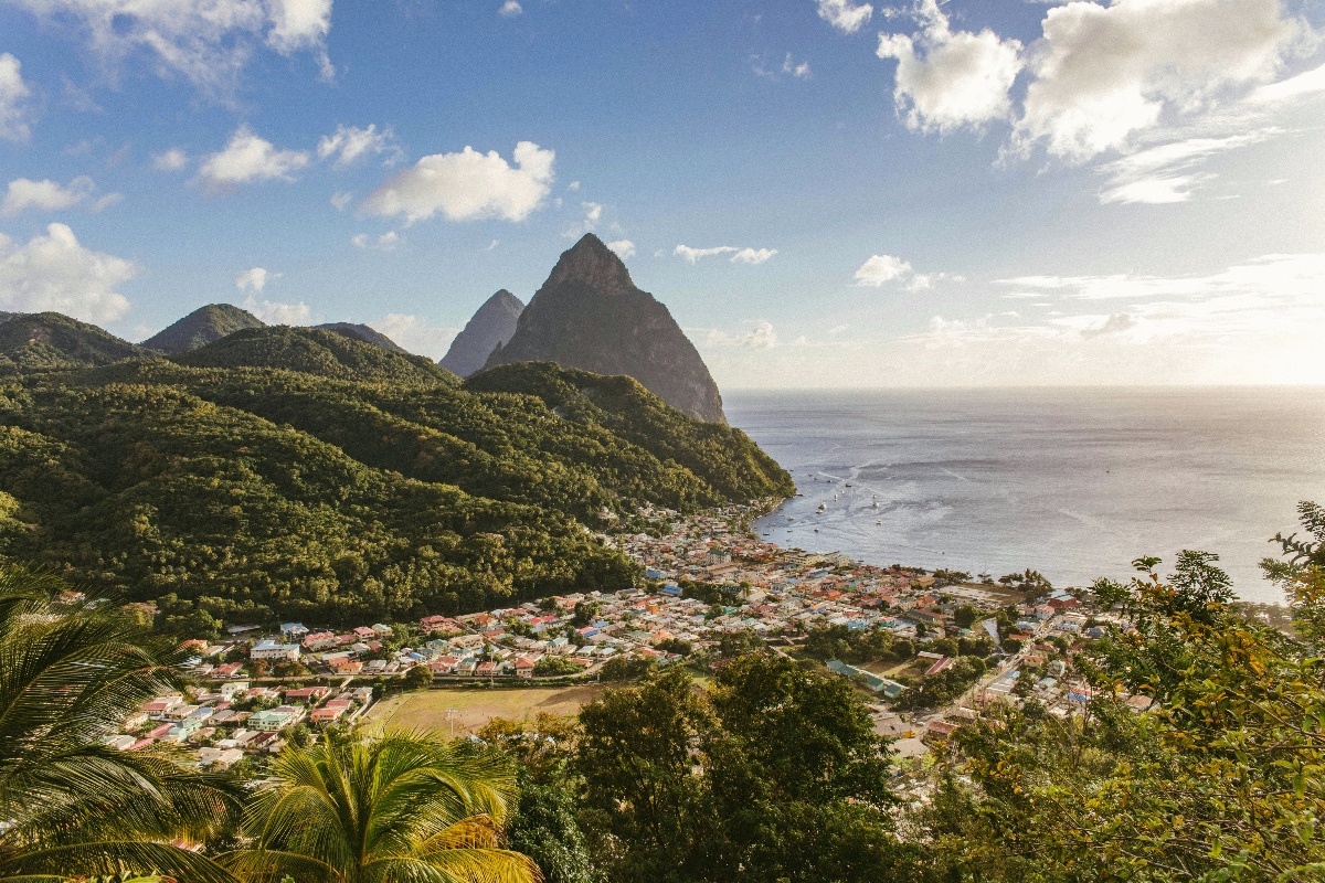 Two tall volcanic peaks rise dramatically from densely forested mountains above a coastal town with red-roofed buildings nestled along a bay. The town sits between the green mountainous terrain and the Caribbean Sea, where several boats are anchored in the calm blue water under partly cloudy skies.