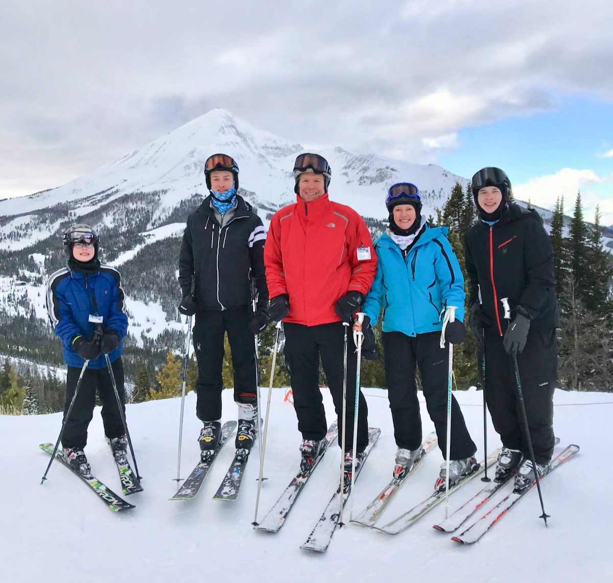 Five skiers stand in a line on a snow-covered slope, wearing helmets, goggles, and winter ski gear in various colors including blue, black, and red. Behind them rises a snow-covered mountain peak with forested slopes beneath a partly cloudy sky.