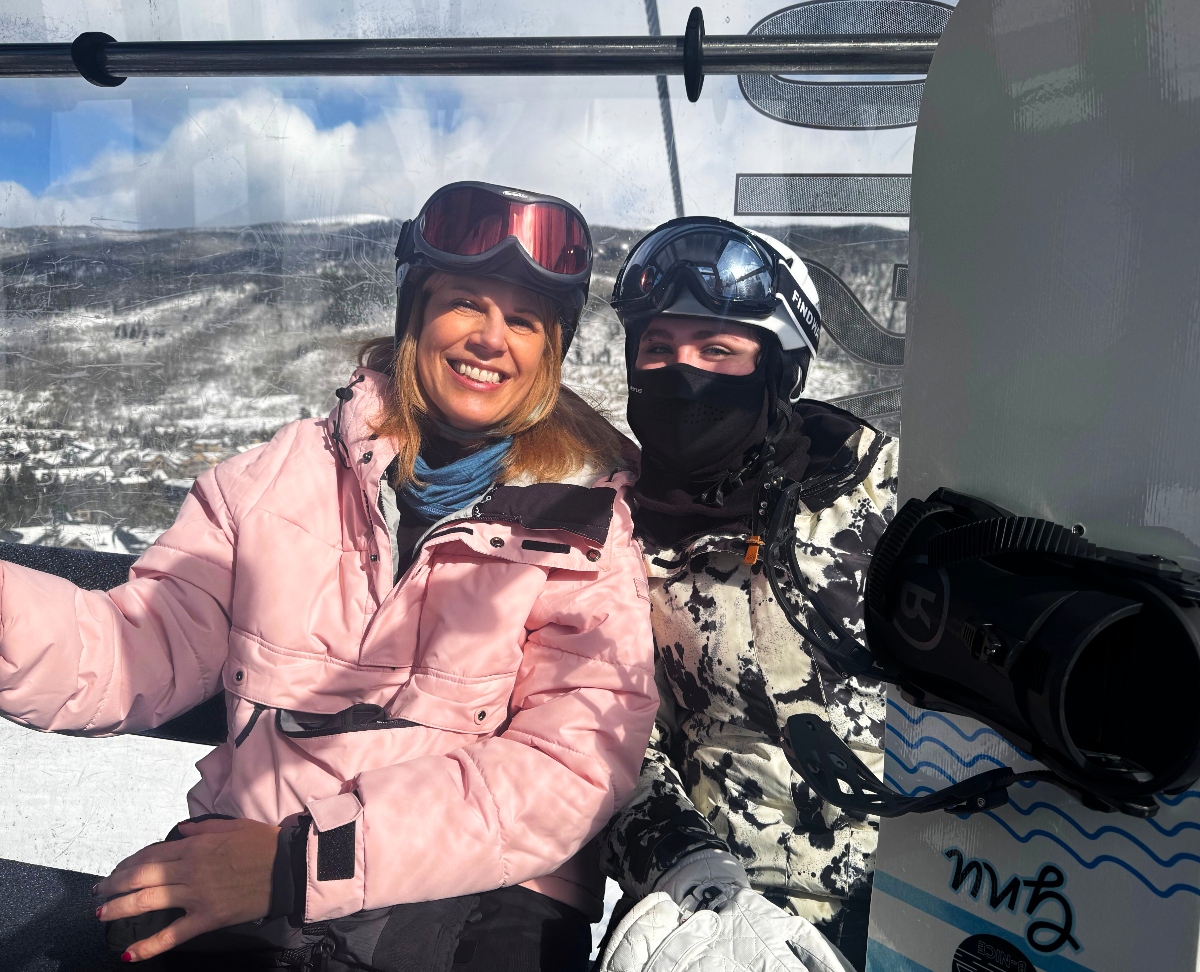Two people ride a ski lift together with snowboards, one wearing a pink ski jacket with goggles and smiling, the other in black winter gear with a face covering. Behind them stretches a snow-covered mountain landscape with forested slopes under partly cloudy skies.