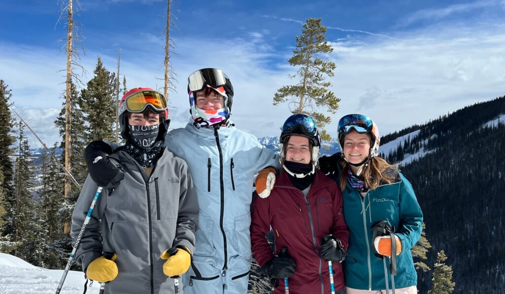 Four skiers wearing helmets and goggles stand together on a snow-covered mountain slope, holding ski poles and dressed in winter jackets of various colors including gray, burgundy, and teal. Behind them, a mountain landscape features evergreen trees, bare standing timber, and distant snow-dusted peaks under a partly cloudy blue sky.