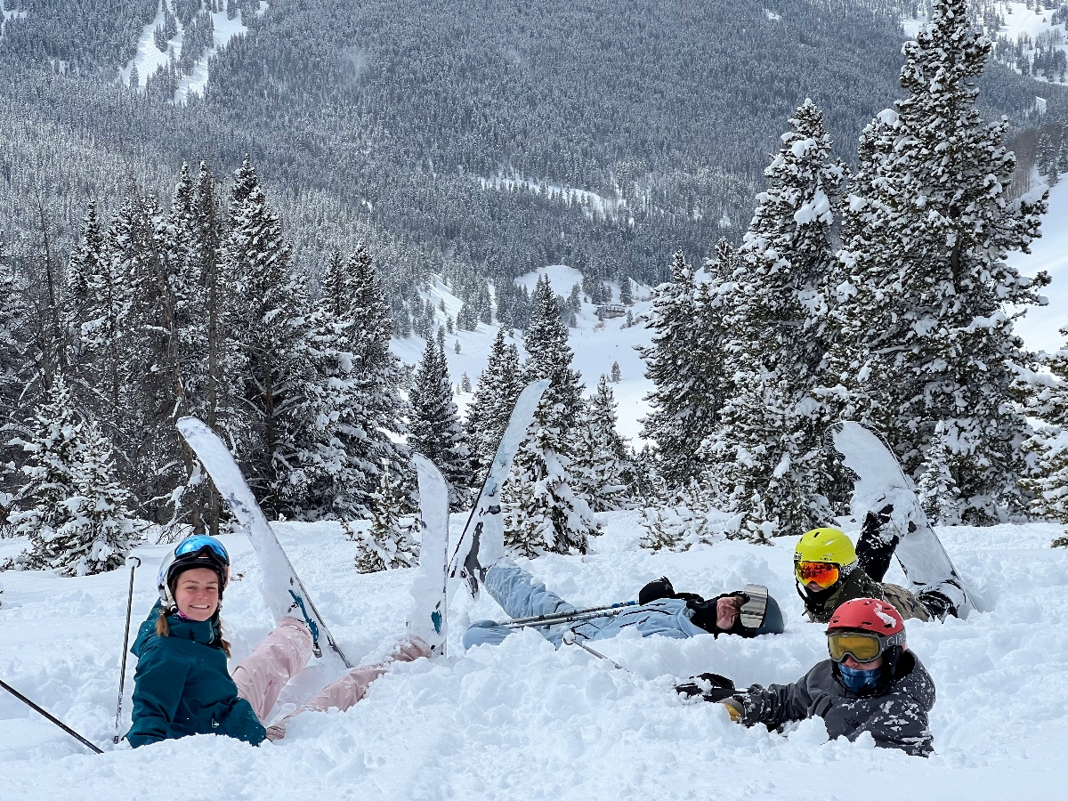 Four skiers in helmets and goggles sit partially buried in deep powder snow with their skis planted upright around them on a mountain slope. Snow-covered evergreen trees frame the foreground while a densely forested mountainside extends into the background under overcast winter conditions.