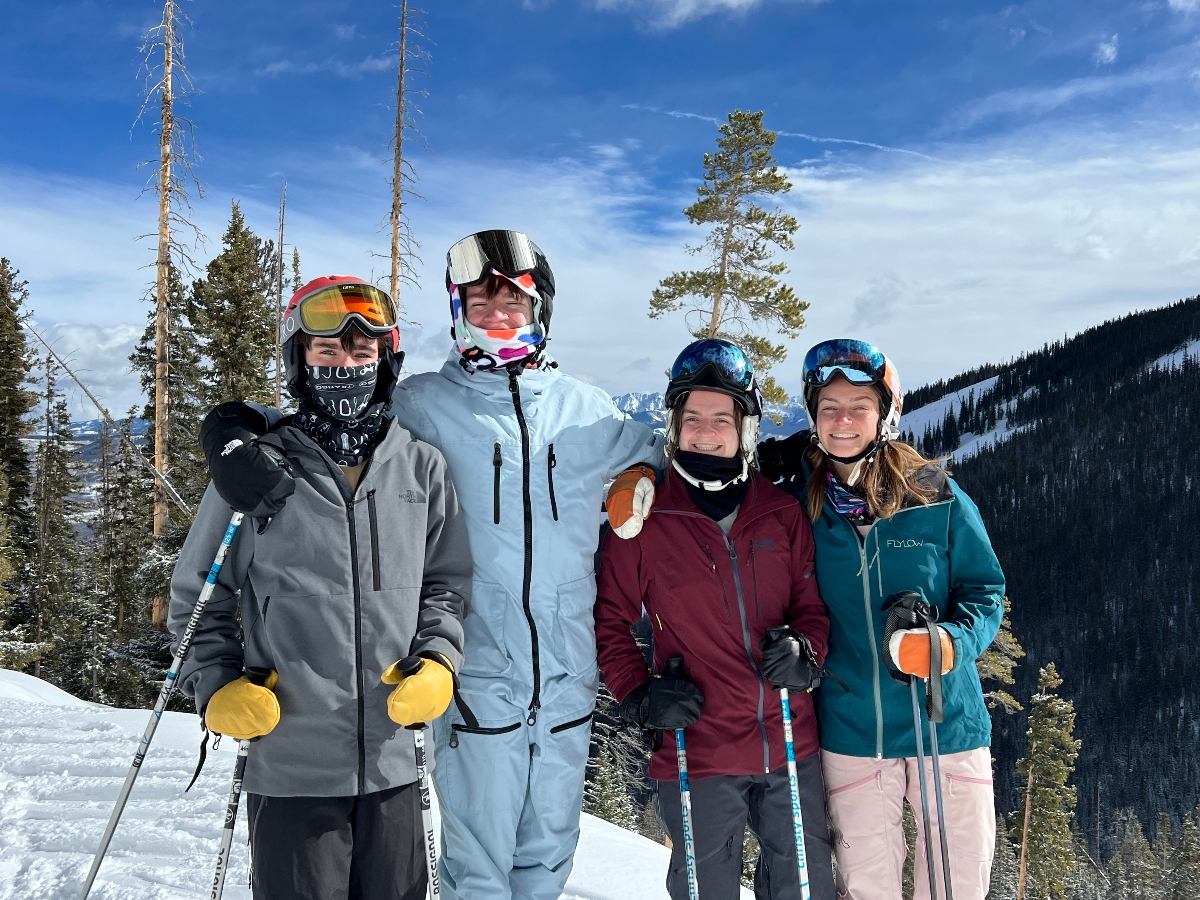 Four skiers wearing helmets and goggles stand together on a snow-covered mountain slope, holding ski poles and dressed in winter jackets of various colors including gray, burgundy, and teal. Behind them, a mountain landscape features evergreen trees, bare standing timber, and distant snow-dusted peaks under a partly cloudy blue sky.