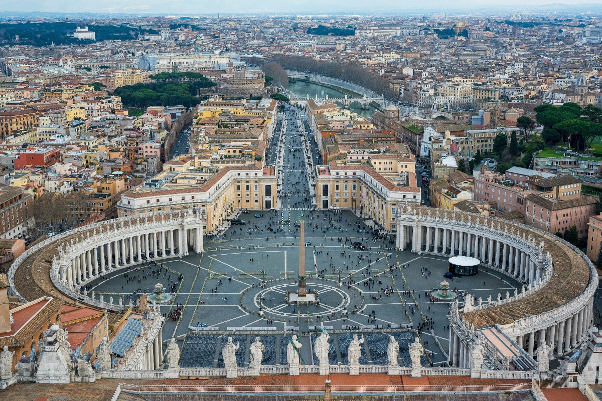 An aerial view looking down from St. Peter's Basilica onto the vast elliptical St. Peter's Square, with its central Egyptian obelisk, two fountains, and geometric paving patterns, enclosed by Bernini's sweeping curved colonnades topped with rows of statues. Beyond the square, the broad Via della Conciliazione extends toward the Tiber River, with the circular Castel Sant'Angelo visible in the middle distance and the dense terracotta-roofed cityscape of Rome spreading to the horizon.