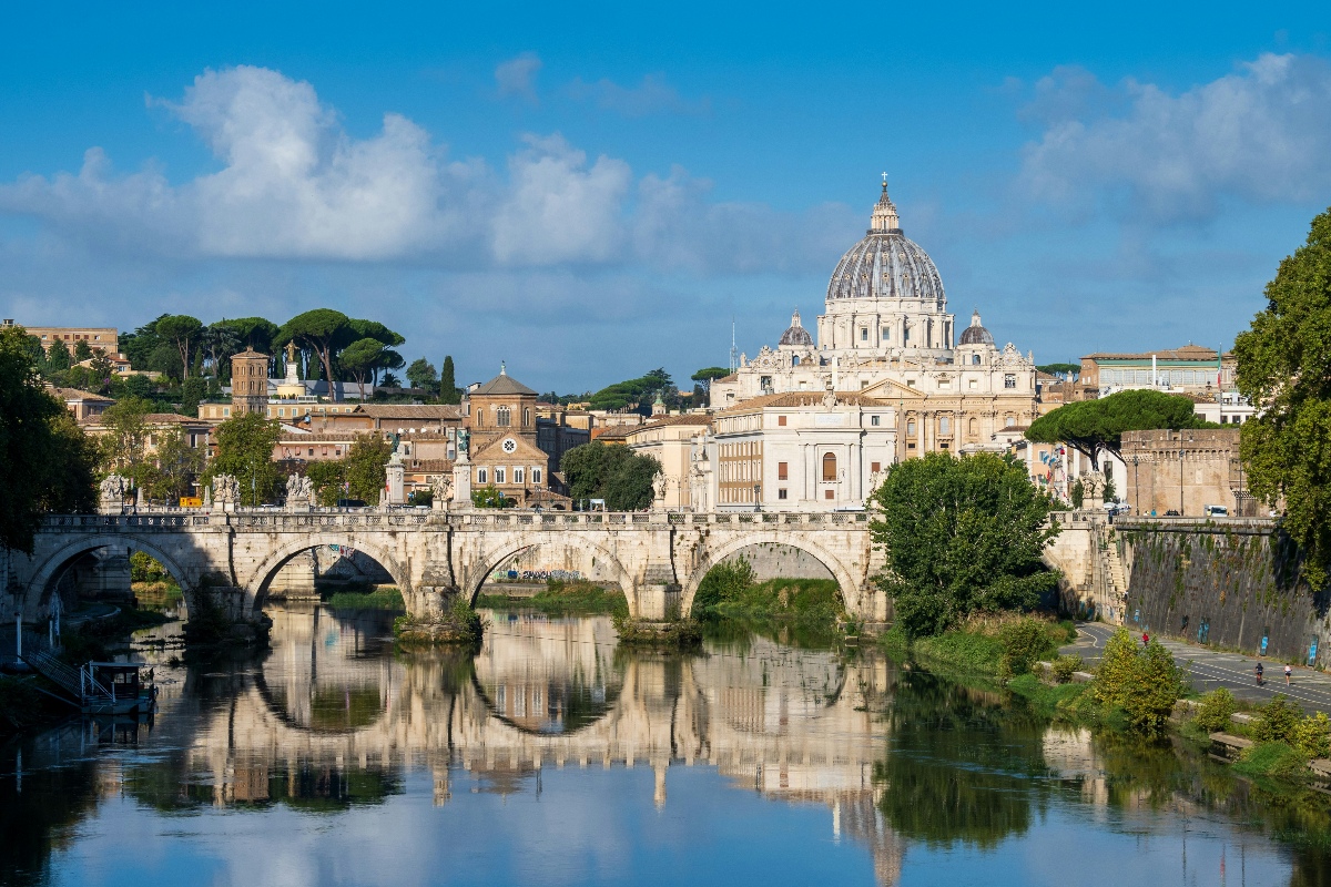 A stone arched bridge spans a calm river, reflecting in the water below. In the background, a large domed cathedral rises above historic buildings and trees under a blue sky with scattered clouds.