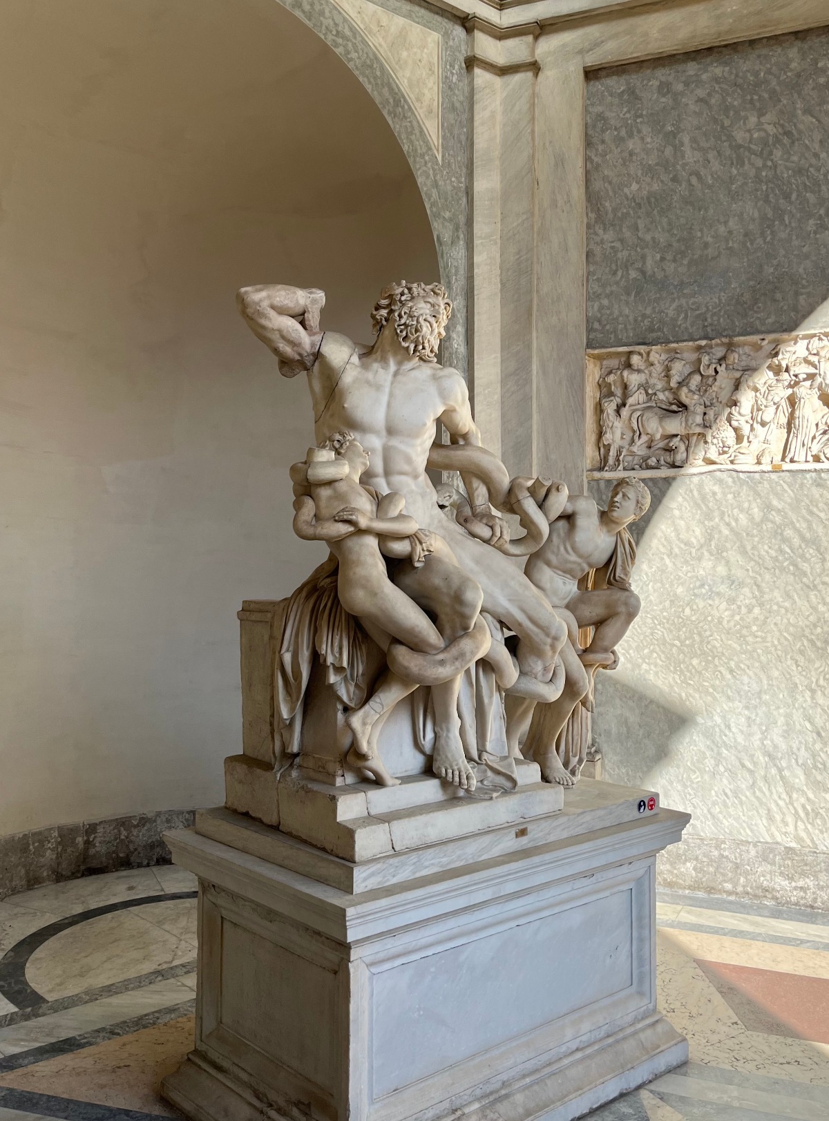 A large white marble sculpture group depicting a bearded man and two younger male figures in dramatic poses, their bodies entwined with serpents, displayed on a rectangular stone pedestal in a museum gallery. The central figure strains upward with one arm raised, his musculature rendered in dynamic detail, set against an arched alcove with decorative stone relief panels visible in the background.
