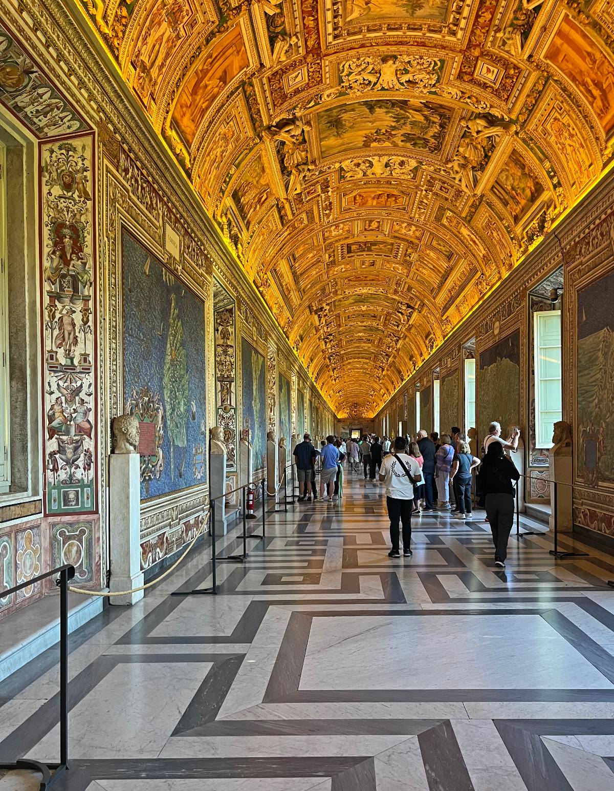 A long, ornate gallery corridor with elaborately painted and gilded barrel-vaulted ceiling stretching into the distance, flanked by large fresco-painted walls depicting maps and landscapes, with classical busts on pedestals along the left side. Groups of visitors walk along the geometric patterned marble floor of cream, gray, and dark stone as they view the artwork lining both walls.