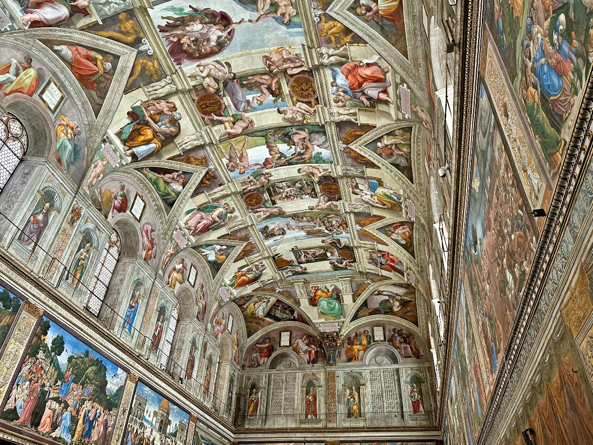 An upward-angled view of the Sistine Chapel's barrel-vaulted ceiling, covered entirely in Michelangelo's frescoes depicting scenes from Genesis, with painted architectural frameworks dividing the surface into panels filled with biblical figures in vibrant blues, reds, greens, and earth tones. The chapel's side walls are also visible, lined with large-scale Renaissance frescoes framed by ornate gilded borders, with arched windows punctuating the upper registers on the left side.