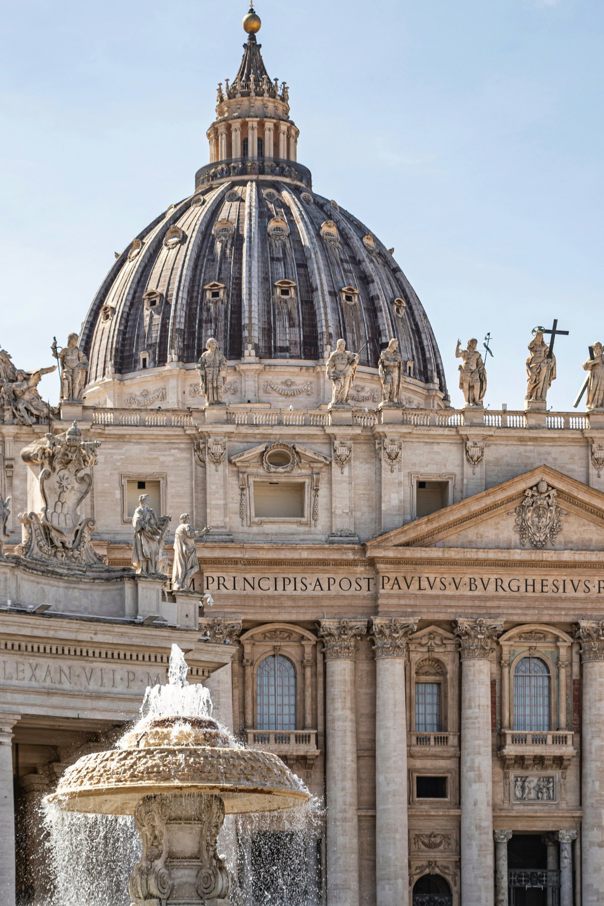A close-up view of the facade and dome of St. Peter's Basilica in Vatican City, showing the ornate travertine stone exterior with large columns, arched windows, and the Latin inscription "PRINCIPIS APOST PAVLVS V BVRGHESIUS" carved across the entablature, with a row of large white marble statues of saints lining the roofline against a pale blue sky. A baroque stone fountain with cascading water is visible in the foreground at the lower left of the frame.