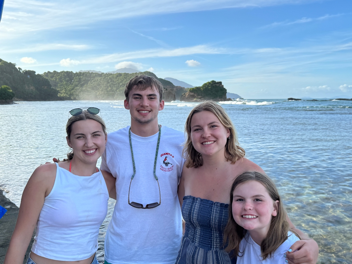 Four young people smile together for a group photo on a scenic coastline, with the young man in the center wearing a white t-shirt and sunglasses around his neck, flanked by three young women in summer clothing. Behind them stretches a beautiful coastal landscape featuring calm blue-green waters, forested hills and rocky outcroppings, with mountains visible in the distance under a partly cloudy sky.