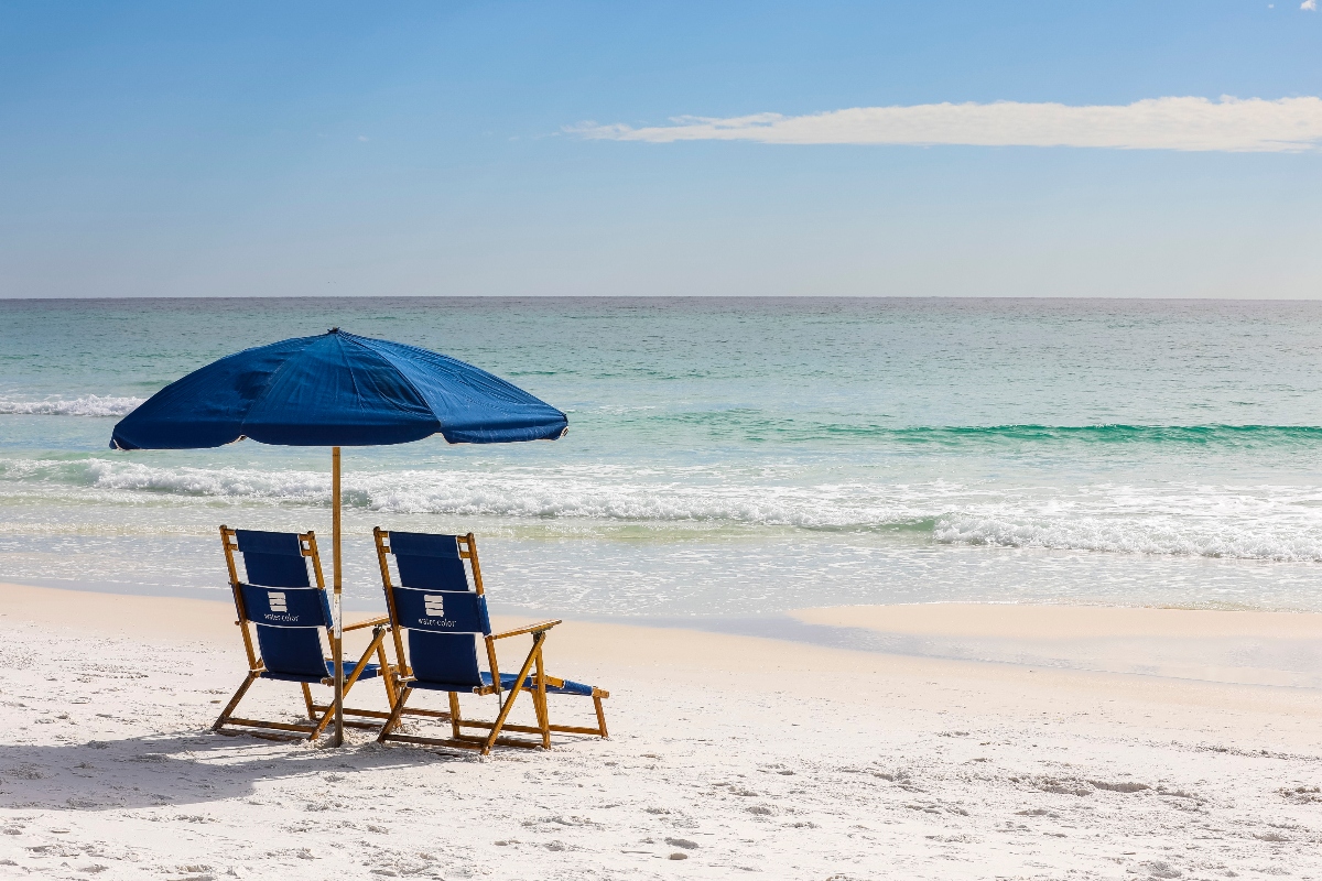 Two deep blue beach chairs and umbrellas sit side by side facing the clear aqua-green water on white sand in late afternoon sunlight. The backs of the chairs feature the logo for WaterColor Inn & Resort. 