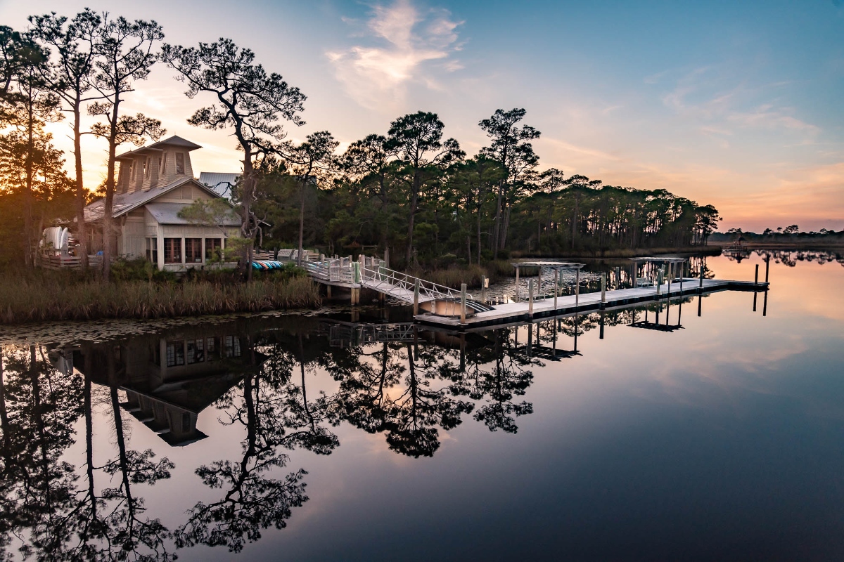 A waterfront boathouse with a cupola sits among pine trees on the shore of a calm lake, connected to a wooden dock where boats are moored. The still water perfectly mirrors the house, trees, dock, and sunset sky with its pink and blue clouds, creating a symmetrical reflection across the scene.