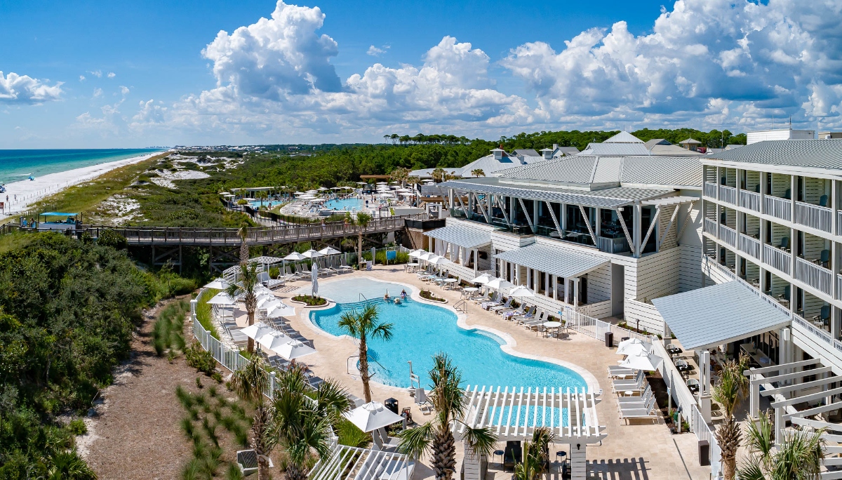 An aerial view shows a beachfront resort complex featuring a large curved swimming pool surrounded by white lounge chairs and palm trees, with multi-story white buildings extending along the property. The resort sits adjacent to a pristine beach and turquoise ocean, connected by wooden boardwalks and surrounded by lush green vegetation under a partly cloudy blue sky.