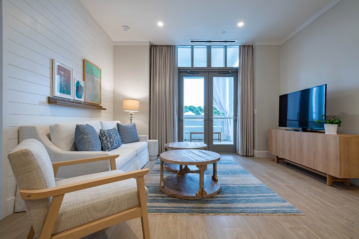 This modern hotel suite living room features a white sectional sofa with blue accent pillows positioned against a white shiplap accent wall, alongside a wooden armchair and round wooden coffee table placed on a blue area rug. The space includes a flat-screen TV on a wooden media console, large windows or glass doors leading to an outdoor balcony, and hardwood flooring throughout.